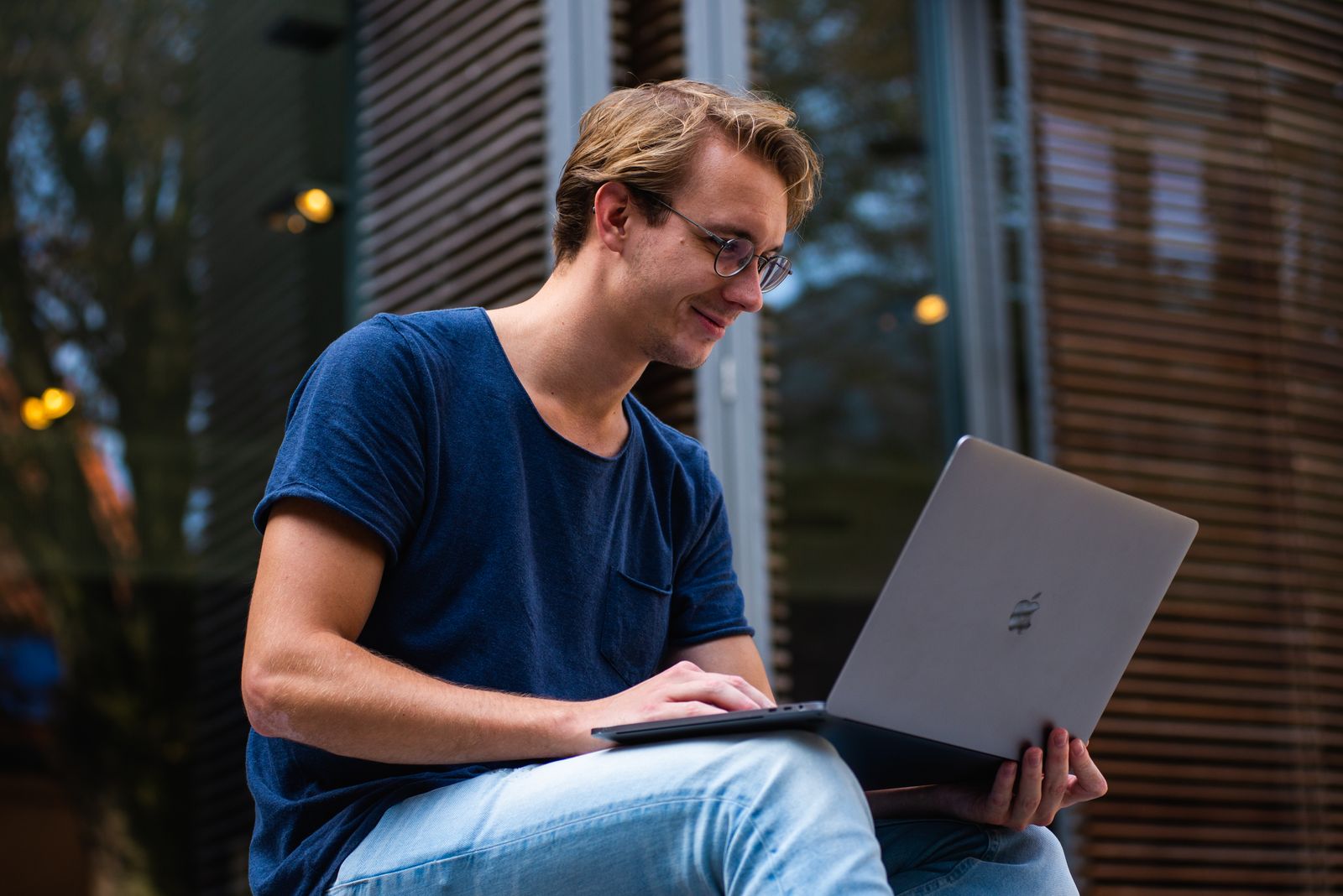 Homem de camiseta azul usa um notebook sentado em uma área externa de um prédio.