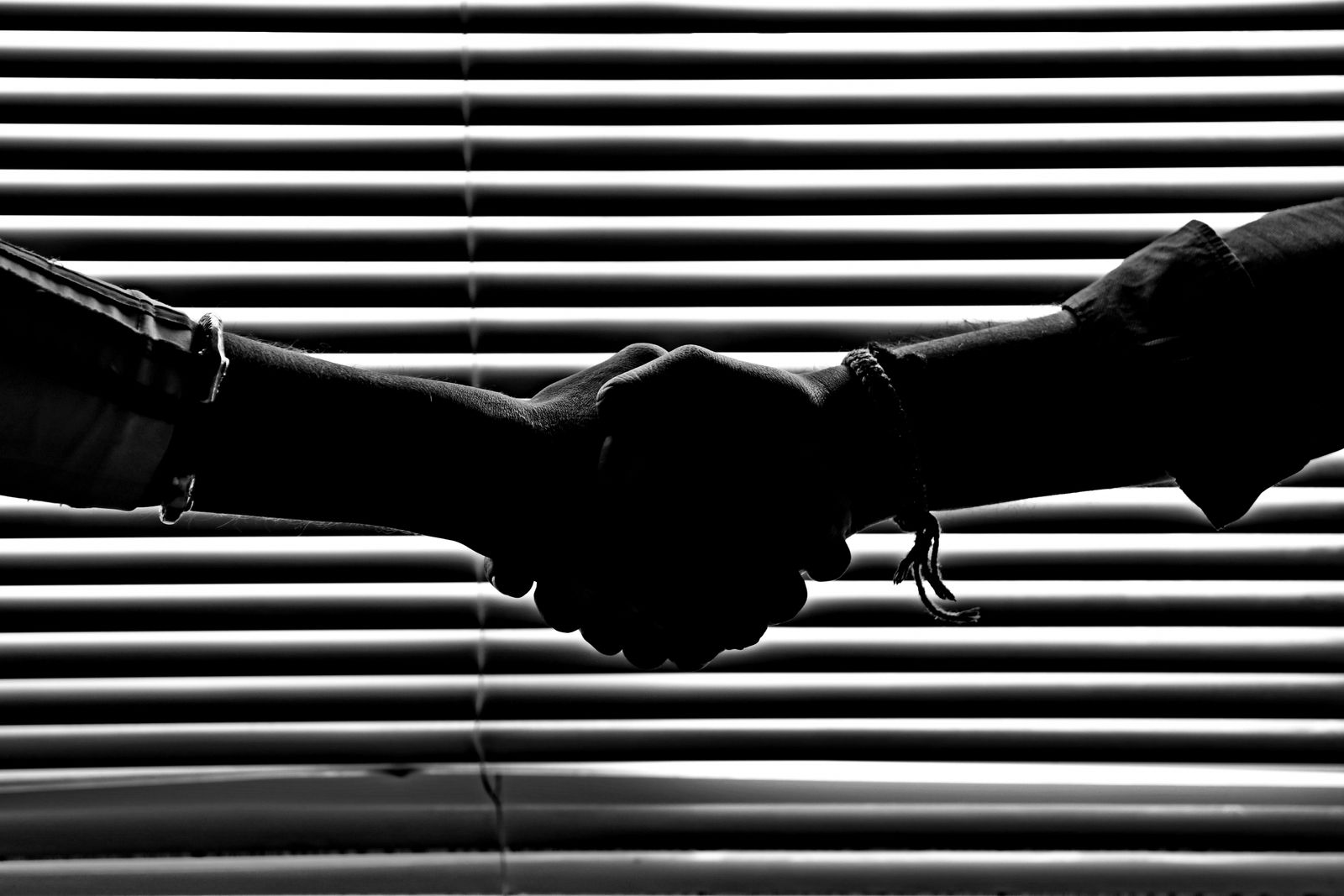 Aperto de mãos em contraluz diante de uma persiana horizontal, em preto e branco.